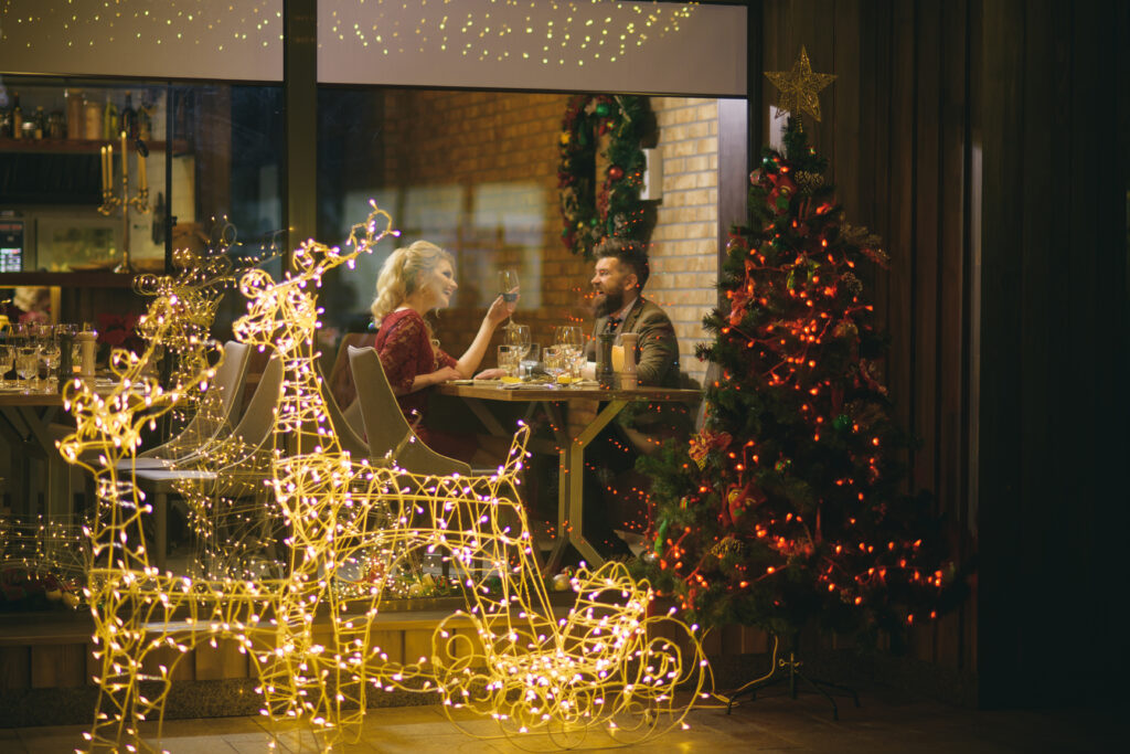 Lampki w kształcie dwóch reniferów ciągnących sanie i udekorowana choinka stoją przed szybą. Za szybą znajduje się wnętrze restauracji. Kobieta i mężczyzna siedzą przy stole i jedzą kolację.
