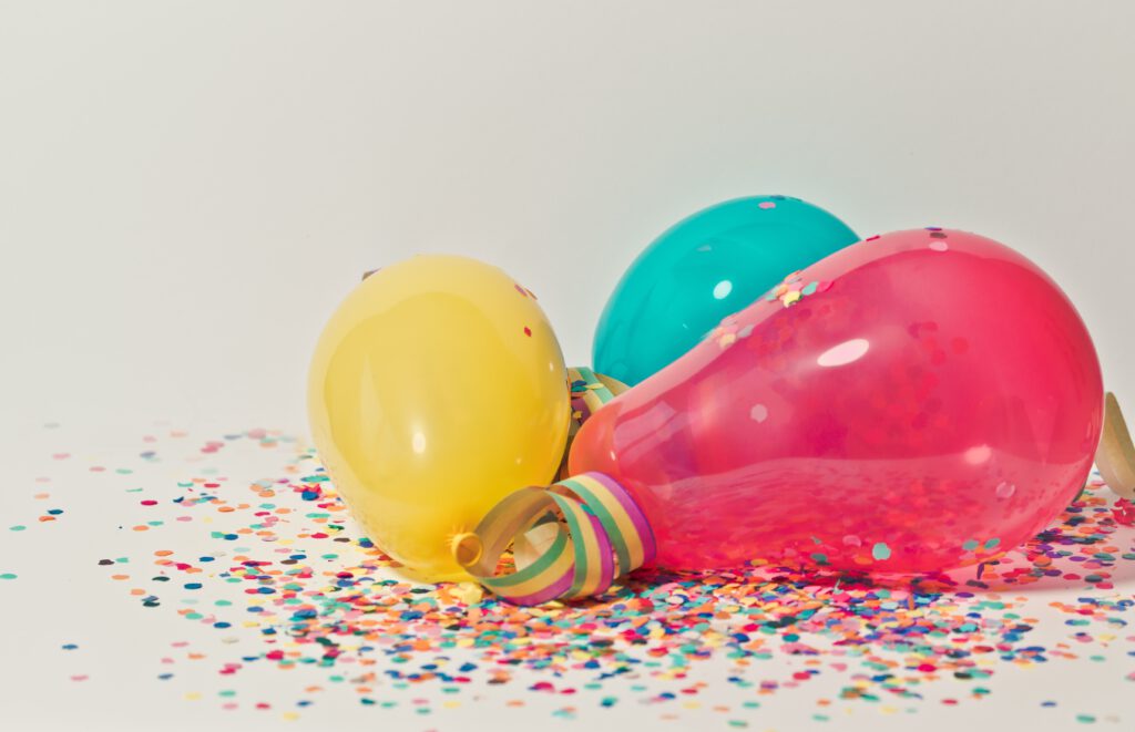 Różowy, żółty i niebieski balon leżą na białej podłodze. Dookoła leży confetti. Całość jest na białym tle,
