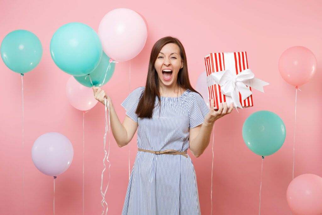 Kobieta stoi na różowym tle. Za nią znajdują się balony. W prawej ręce trzyma niebieskie i różowe balony, a w lewej - prezent.