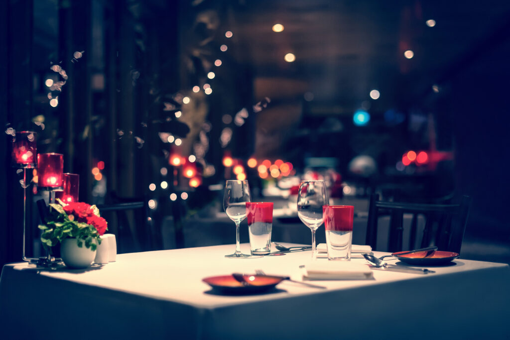 Stół zasłany białym obrusem, a na blacie elegancka zastawa. W tle zamglone wnętrze restauracji.