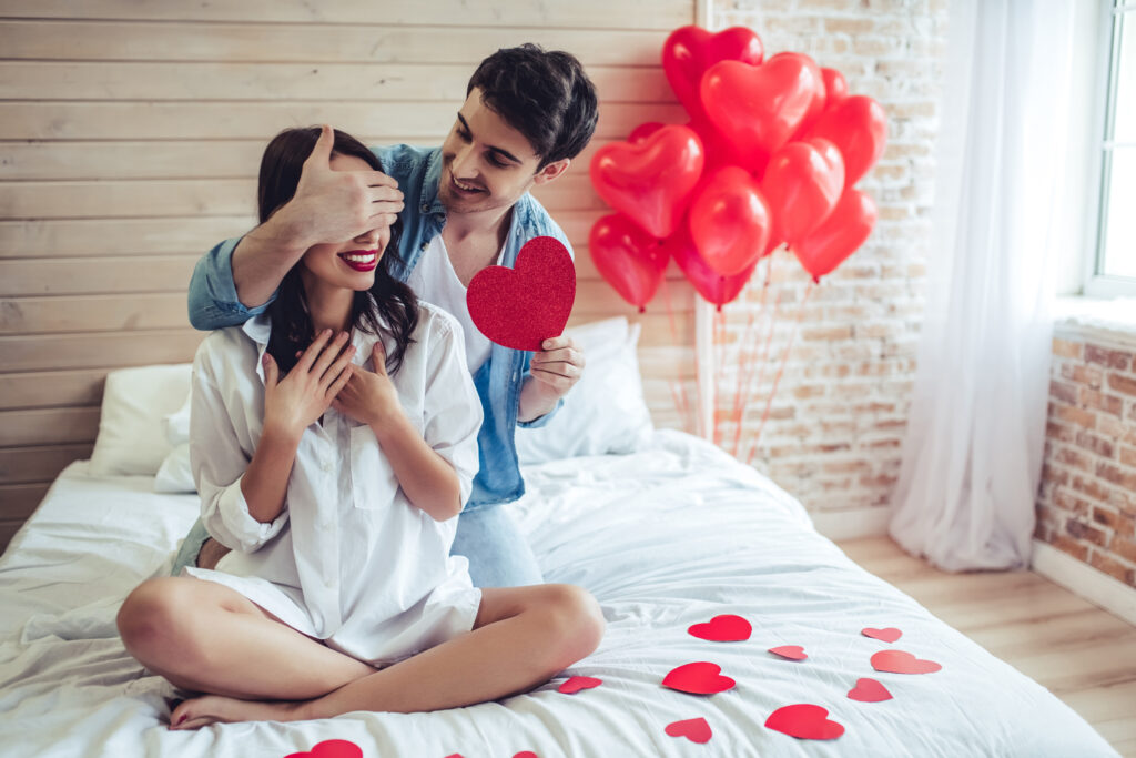 Mężczyzna zasłania kobiecie oczy i podaje papierowe serce. Oboje siedzą na łóżku. W tle balony w kształcie serca, drewniane i murowane ściany pokoju i kawałek okna.