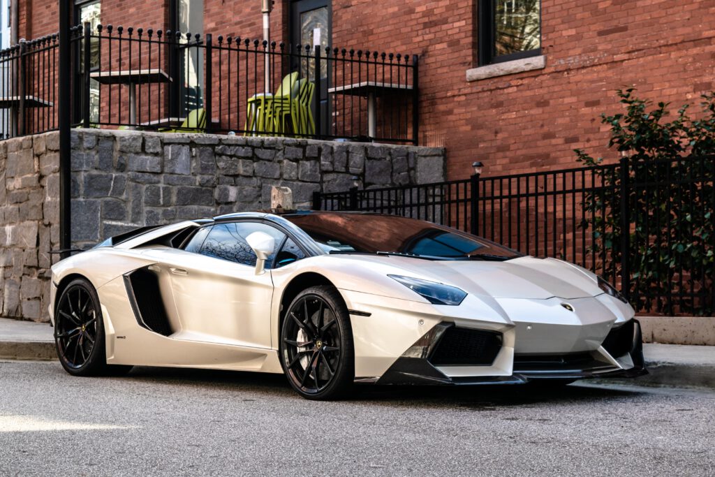 Białe lamborghini stoi na ulicy. W tle murowany dom i metalowe bramy.