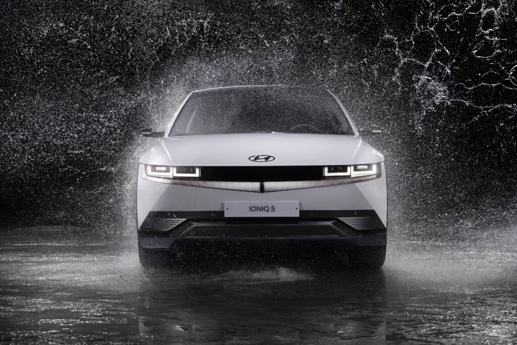 Biały samochód Hyundai IONIQ 5 stoi przodem na drodze. Dookoła rozbryzguje woda.