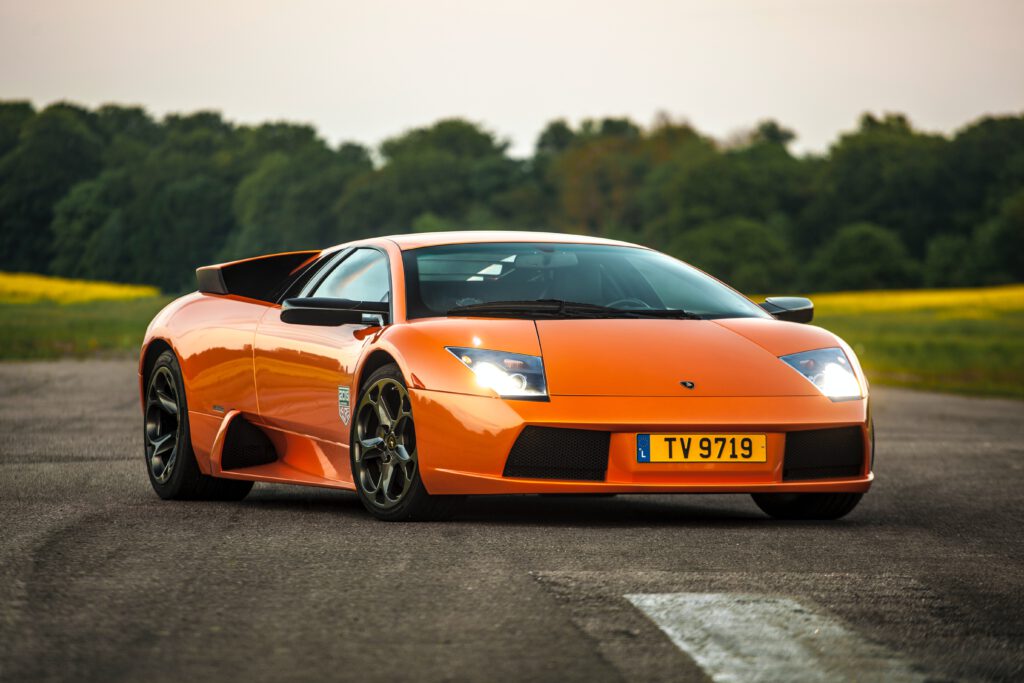 Pomarańczowe lamborghini stoi na drodze. W tle niebo, lasy i pola.