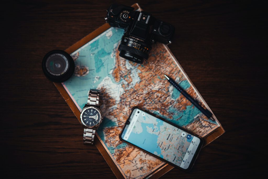 Na stole leży mapa Europy. Na niej telefon z wyświetloną mapą Europy, aparat fotograficzny, niebieski długopis oraz zegarek na rękę.