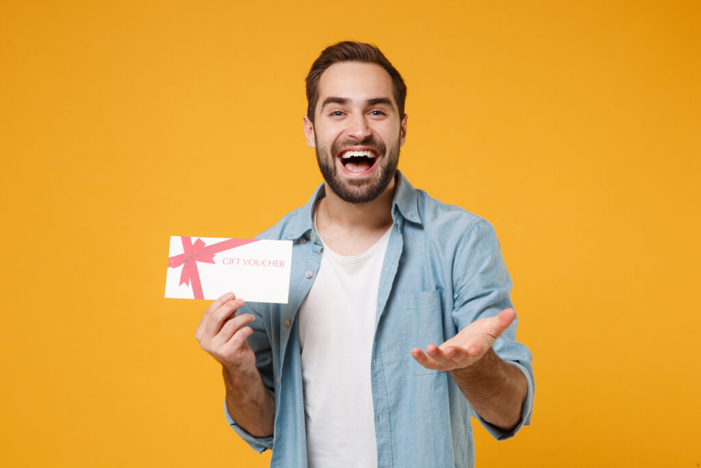 Mężczyzna w białej koszulce i niebieskiej koszuli stoi na żółtym tle. Uśmiecha się, a w prawej dłoni trzyma Voucher. Lewą rękę wyciąga do przodu.