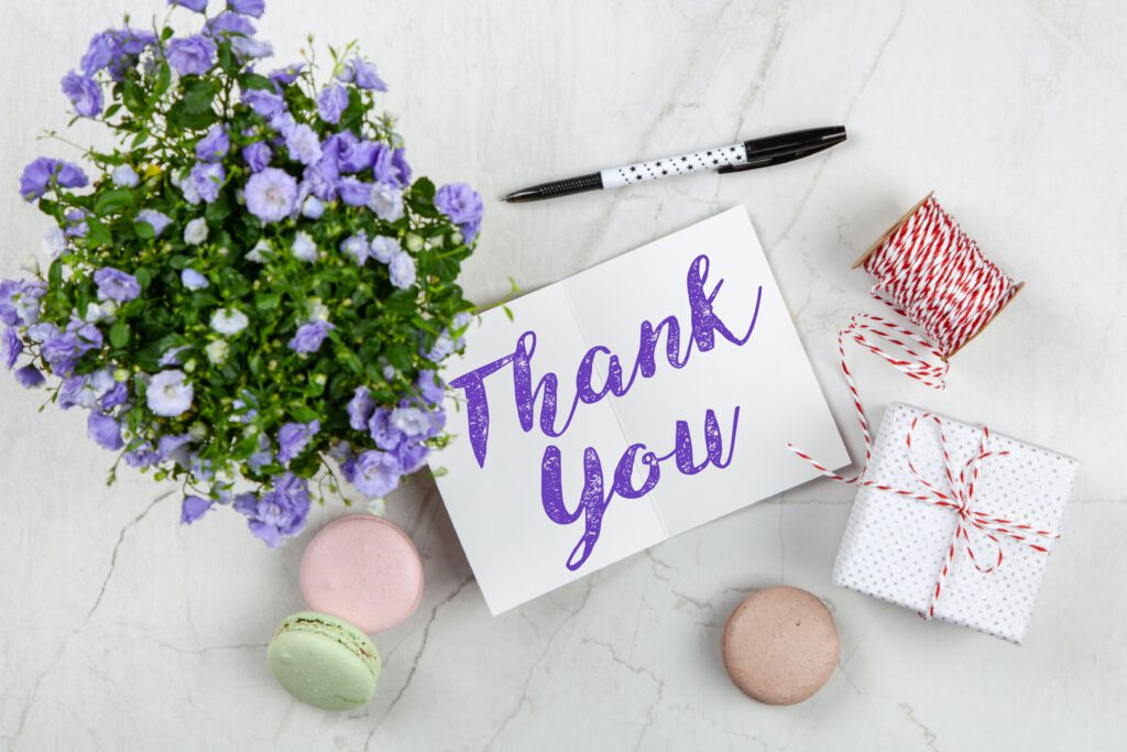 Biała kartka z fioletowym napisem ,,thank you". Obok stoi bukiet kwiatów, leżą ciasteczka, stoi pudełko i leży sznurek.