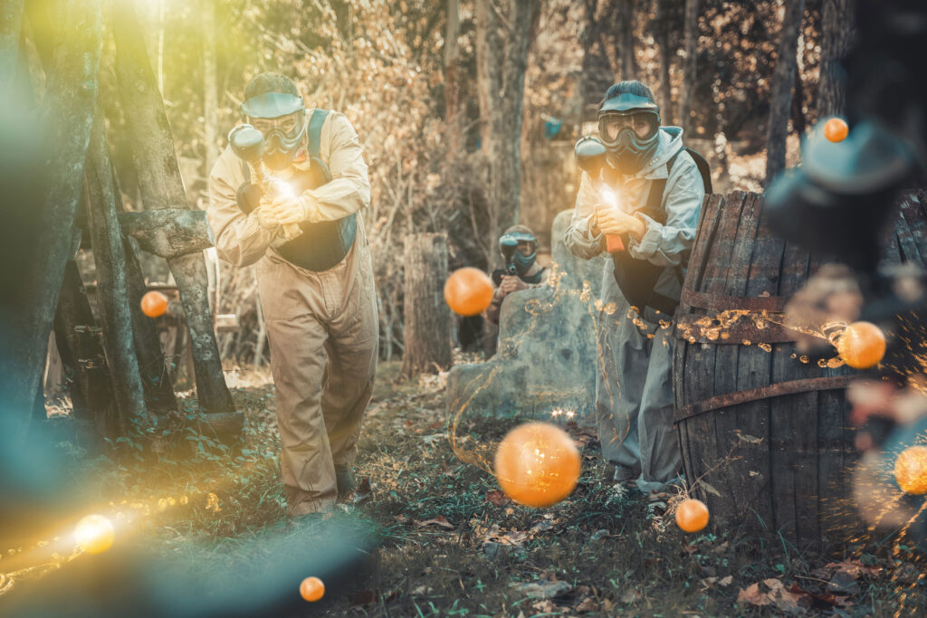 Mężczyzna i kobieta grają w paintballa. Z tyłu za kamieniem klęka mężczyzna. W tle drzewa.