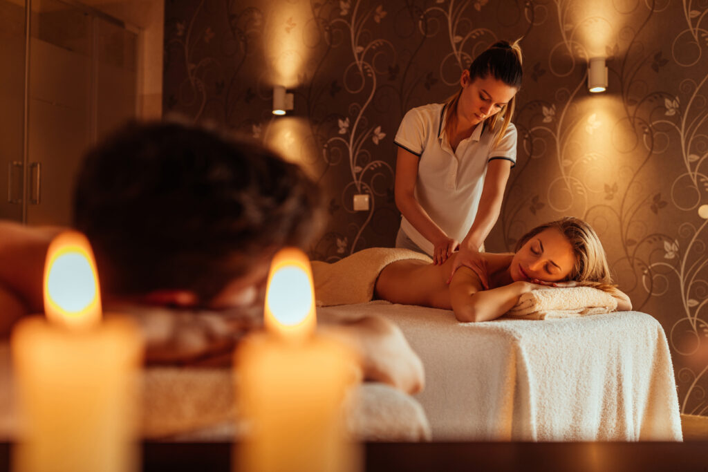 Kobieta i mężczyzna leżą na stołach do masażu w salonie SPA. Masażystka masuje kobietę. W tle świece, brązowa tapeta i lampy.
