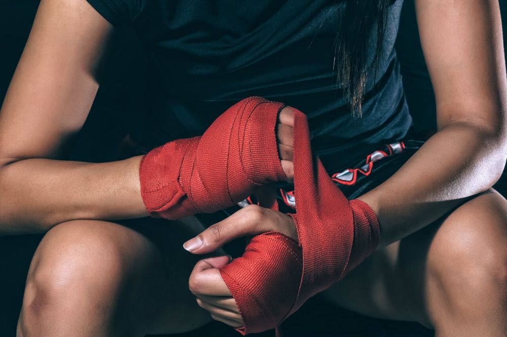 Osoba przygotowująca się do treningu sztuk walki owija dłonie czerwonymi bandażami bokserskimi – symbol determinacji i siły, idealny kadr promujący voucher na sporty walki.