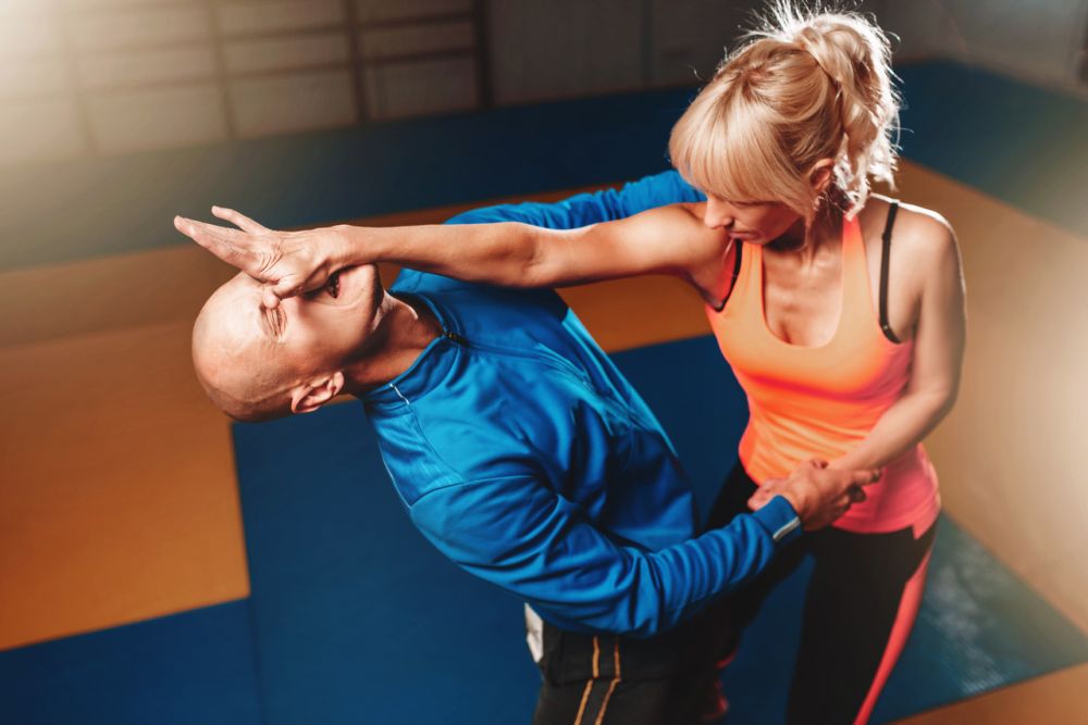 Kobieta w sportowym stroju ćwiczy technikę samoobrony, blokując i odpychając przeciwnika na macie treningowej podczas zajęć ze sztuk walki.