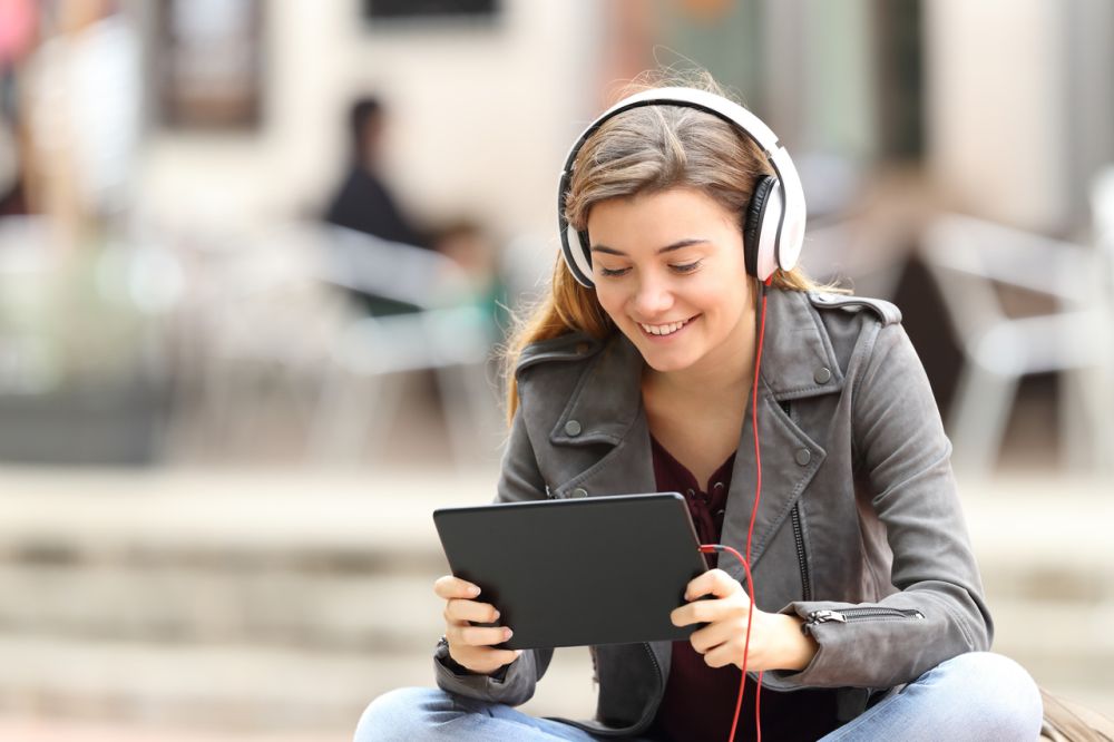 Uśmiechnięta młoda kobieta ucząca się języka obcego online – słucha lekcji na słuchawkach i korzysta z tabletu w plenerze, nowoczesna nauka w przyjaznej atmosferze.