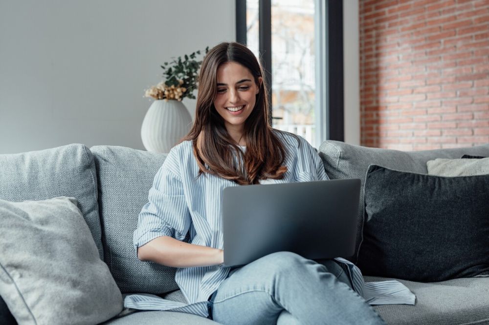 Kobieta ucząca się języka obcego online na laptopie w domowym zaciszu – uśmiechnięta, skoncentrowana na ekranie, wygodna nauka języka z kanapy.